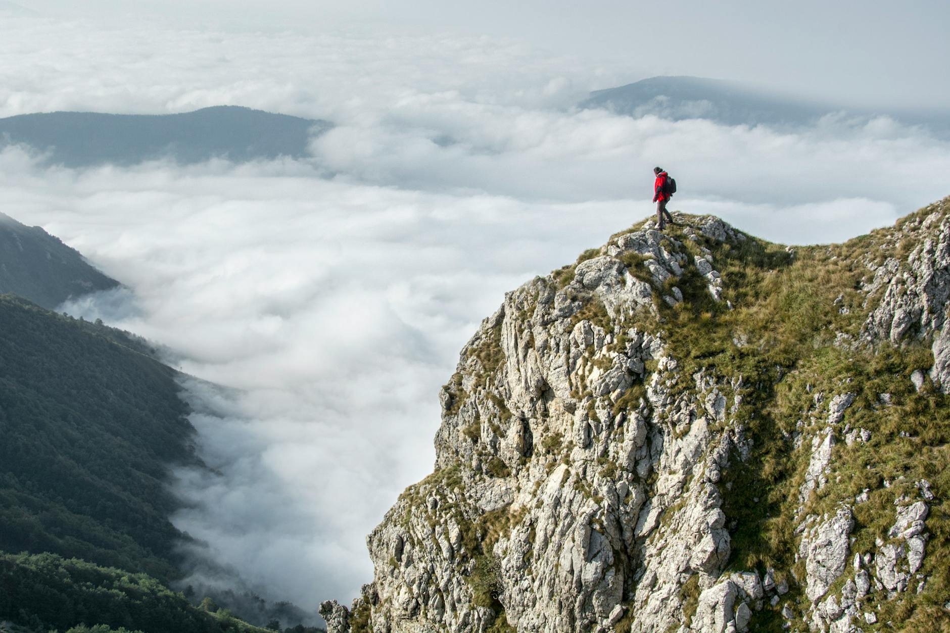 A man wearing an orange jacket and carrying a backpack stands at the top of a mountain, looking out at clouds and green mountain peaks, lower down.